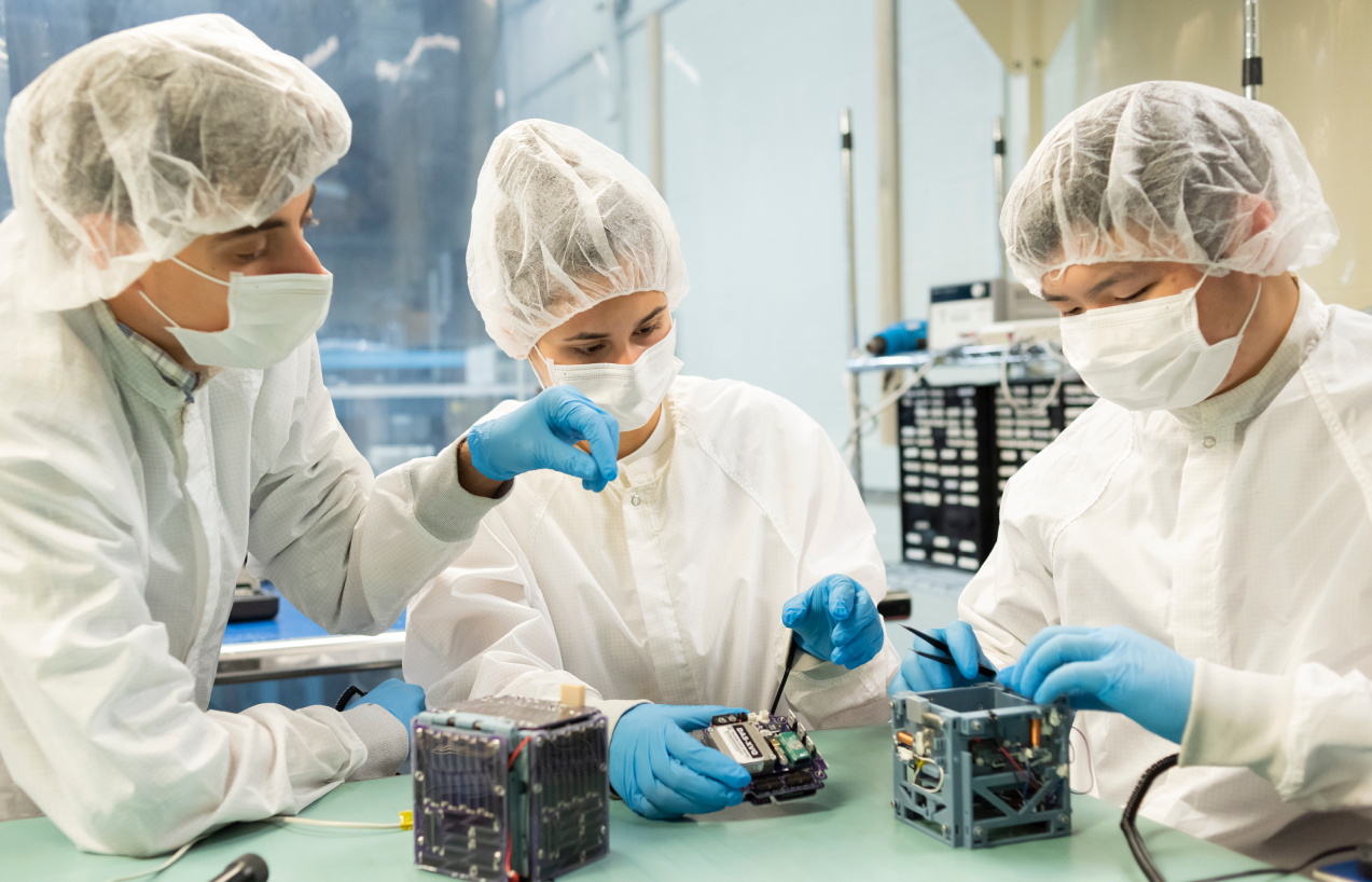 Action shot of Alpha team members Josh, Ally, and Andy assembling the CubeSat (at right) based on its prototype (at left).