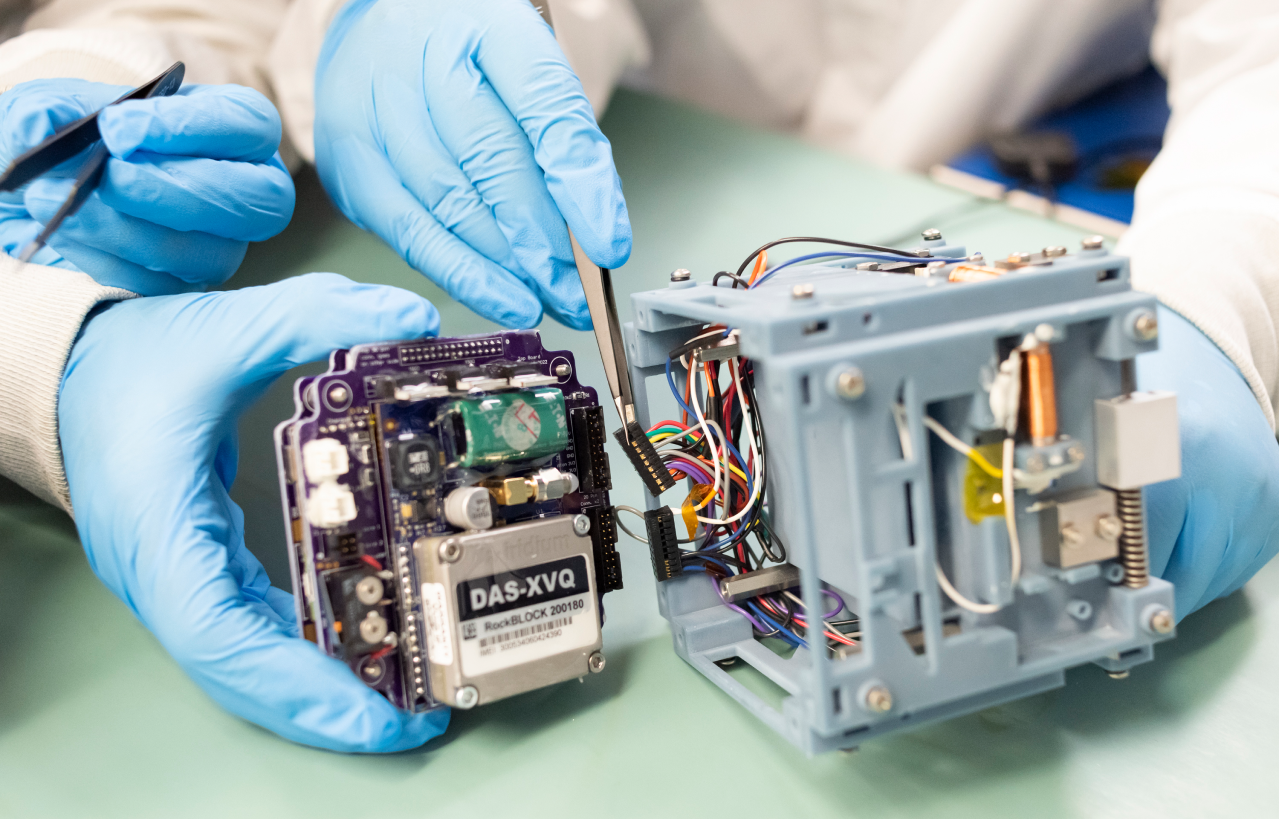 Action shot of the electronics stack being integrated into the CubeSat. The RockBLOCK is the shiny metal box at left; one of the copper magnetorquers can be seen on the CubeSat’s right side.